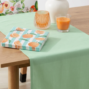 Mediterrane Tischdekoration mit grünem Tischläufer, Zitrus-Kissen, Orangen-Servietten, Windlicht und Kerze für sommerliches Wohnambiente.